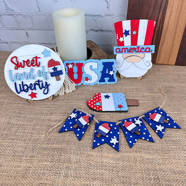 Introducing the "Patriotic - Tiered Tray Kit" by Janet's Craft Corner, a delightful DIY kit for patriotic enthusiasts. This set features farmhouse-style decor on a burlap surface and includes a "Sweet Land of Liberty" sign, "USA" letters, an American flag-hat gnome, boot ornament, and star-spangled banner with wooden elements. A candle in the background adds a warm glow to complete the charming display.