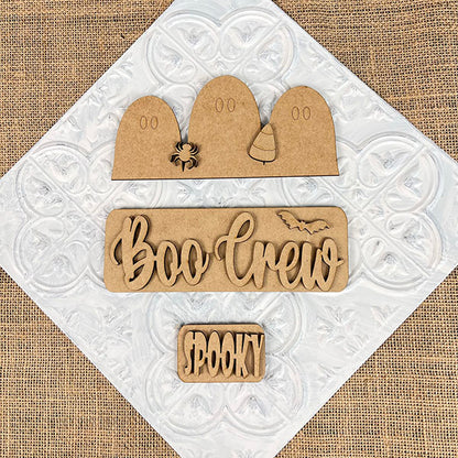 Featuring the "Boo Crew Interchangeable Insert" DIY home decor kit from Janet's Craft Corner, this hand-painted craft set includes wooden ghost shapes, a spider, and a candy corn on a textured background. It also showcases the phrases "Boo Crew" with a bat and "Spooky," making it an ideal interchangeable accessory for adding festive charm to any space.