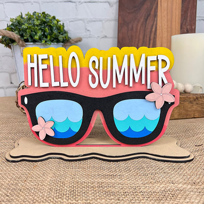 The Hello Summer Sunglasses Shelf Sitter by Janet's Craft Corner showcases oversized sunglasses with blue lenses and pink frames, embellished with pink flowers, for a beachy farmhouse decor vibe. The text "Hello Summer" is elegantly displayed above the sunglasses as it sits on a table against a brick wall backdrop with a candle nearby.