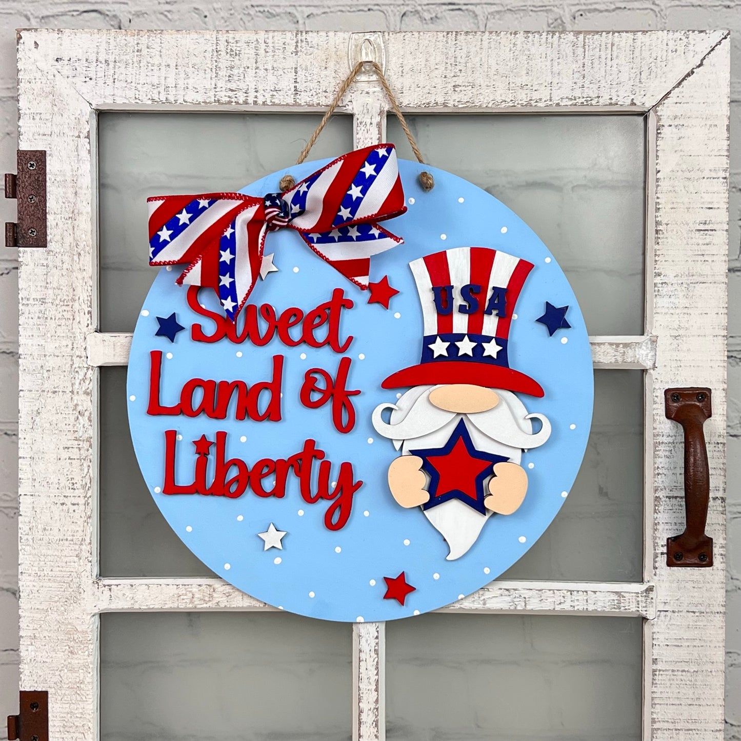 The Sweet Land of Liberty Door Hanger by Janet's Craft Corner features a gnome wearing a red, white, and blue hat while holding a star. The bold phrase "Sweet Land of Liberty" is written in red. Accentuated with a festive bow and stars, it radiates patriotic charm.