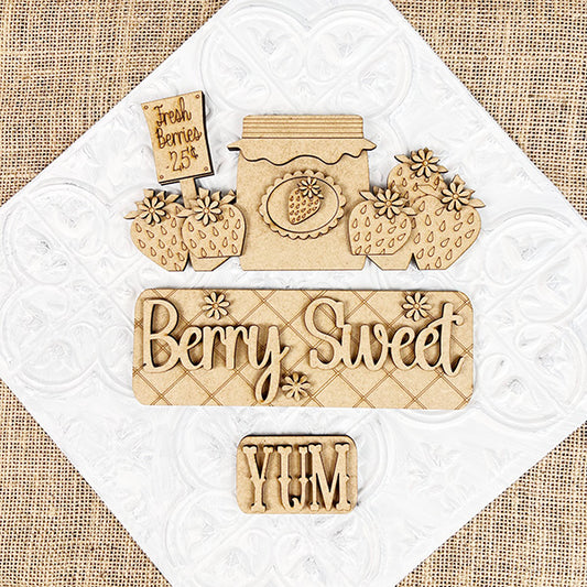 Arranged on a white textured background are wooden cutouts from Janet's Craft Corner, featuring a "Fresh Berries - 25¢" sign, a jam jar, strawberries, "Berry Sweet" text, and a "YUM" piece. This Berry Sweet Interchangeable Insert is part of their DIY home decor kit that's perfect for customizable farmhouse décor on a charming burlap surface.