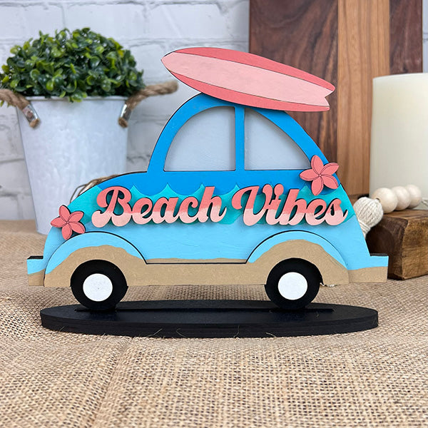 The "Beach Vibes Volkswagen" by Janet's Craft Corner showcases a charming wooden home décor piece, featuring a blue Volkswagen car with a surfboard on top. The side is embellished with "Beach Vibes" in pink letters and accented by pink flowers. It rests on a burlap-covered surface, surrounded by plants and candles, evoking a delightful coastal ambiance.