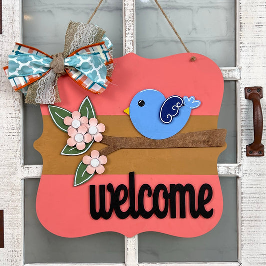 A delightful Bird Welcome Sign from Janet's Craft Corner enhances the door, featuring a blue bird on a branch decorated with pink flowers and green leaves. A vibrant bow in blue, orange, and lace patterns tops this charming home décor piece, with "welcome" prominently displayed underneath.