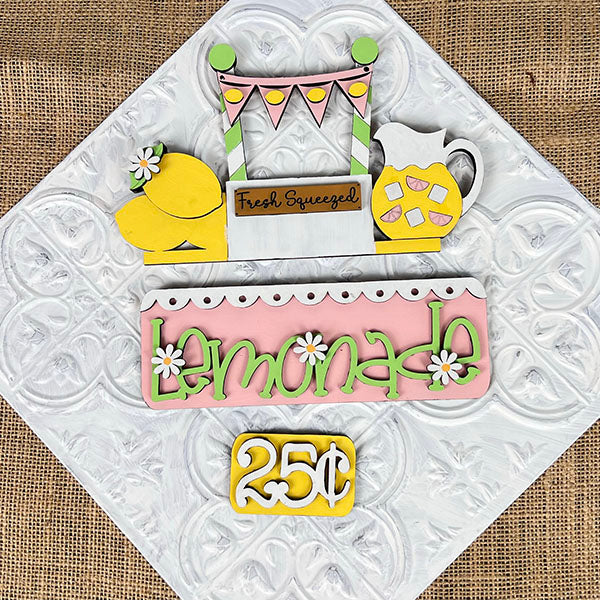 The "Lemonade Interchangeable Insert," part of Janet's Craft Corner DIY home decor kit, features vibrant cutouts of lemons, a pitcher, and a banner. Ideal for enhancing your living space, it sits on a textured white background that mimics embossed tin.