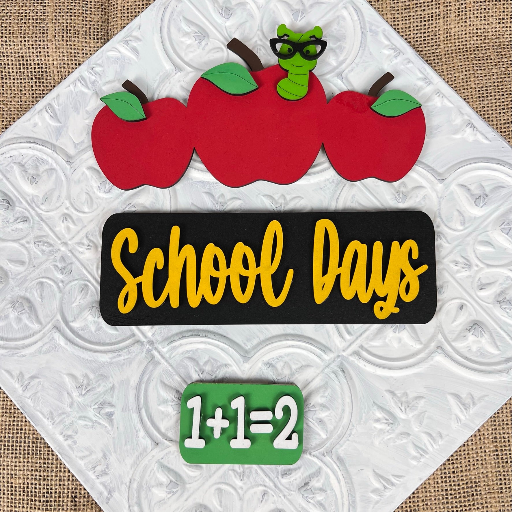 The School Days Interchangeable Insert by Janet's Craft Corner showcases a farmhouse decor sign featuring three apples, one with a quirky green worm wearing glasses. The phrase "School Days" is prominently displayed in yellow against a black background, with "1+1=2" underneath. This piece serves as an ideal addition for interchangeable craft inserts or to enhance your DIY craft kit projects, set against a textured white pattern backdrop.