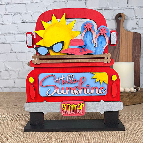 The Hello Sunshine Interchangeable Insert from Janet's Craft Corner is a hand-painted DIY home decor kit featuring a decorative red truck adorned with summer-themed elements: a large sun sporting sunglasses, a pink hat, blue flip-flops, and the phrases "Hello Sunshine" and "Summer" on the back. This charming piece makes an ideal addition to any brick wall backdrop for home decor enthusiasts.