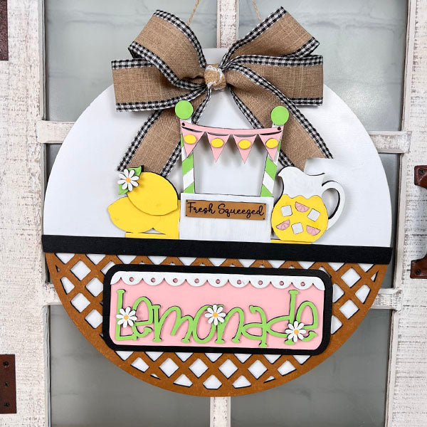 Janet's Craft Corner presents the Lemonade Interchangeable Insert, a delightful round door hanger DIY home decor kit. Featuring a lemonade theme with a pitcher of refreshing lemonade, fresh lemons, and a "Fresh Squeezed" sign, it's topped with a charming burlap and checkered ribbon bow. This hand-painted craft kit is perfect for any season due to its interchangeable insert feature.