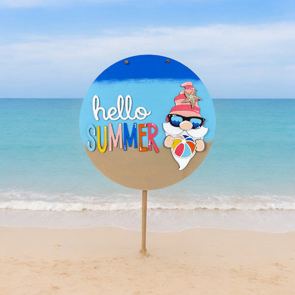 A colorful beach sign features "hello SUMMER" with a character resembling Janet's Craft Corner's Hello Summer Gnome Door Hanger, wearing sunglasses and a starfish-adorned red hat. Perfect for home décor, it stands on a sandy beach with a calm blue sea and sky backdrop.