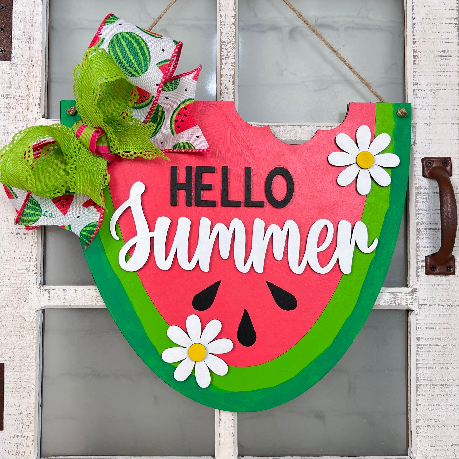 The Hello Summer Watermelon Door Hanger by Janet's Craft Corner features a charming design with a red center, black seeds, and green rind. The phrase "Hello Summer" is beautifully written in white, accompanied by daisy flowers and a green and white bow, making it an ideal piece of summer décor to enhance any wooden backdrop.