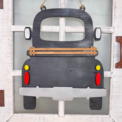 A decorative wooden cutout of Janet's Craft Corner's Antique Truck Interchangeable Base - Painted, seen from the rear with red and yellow taillights, brown side rails, hanging on a white window for charming interchangeable decor.