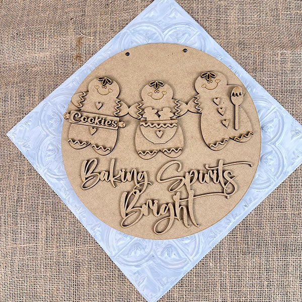 The Baking Spirits Bright Wall Hanger by Janet's Craft Corner is a DIY seasonal home décor kit featuring a wooden plaque with three gingerbread figures and the text "Baking Spirits Bright" on a white textured square over burlap.