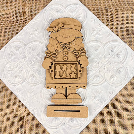 Janet's Craft Corner offers the Mrs Claus Baking Gnome Shelf Sitter, a charming wooden figure donning a hat and holly. Holding a plaque with festive shapes, it's perfect for your holiday display, set on a textured white background on burlap. This DIY seasonal home decor craft kit comes as a set of 1.