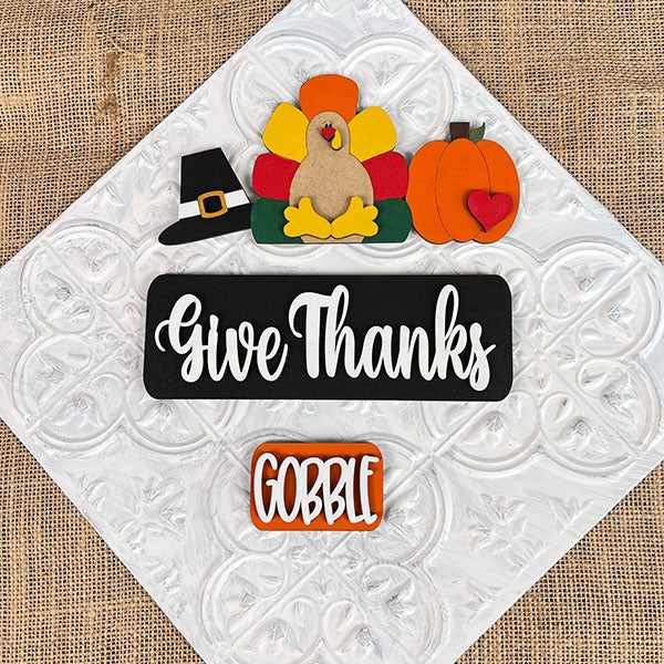 Enhance your home for Thanksgiving with Janet's Craft Corner's Give Thanks Interchangeable Insert - Painted, featuring a textured surface adorned with hand-painted ornaments such as a turkey, a pilgrim hat, and a pumpkin with a heart. Finish off your festive decor with two delightful signs that say "Give Thanks" and "Gobble," perfect for interchangeable craft displays!