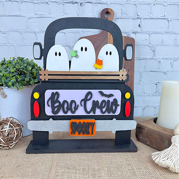 A decorative display showcases a charming trio of ghost figures in the back of a black truck, crafted with Janet's Craft Corner's Boo Crew Interchangeable Insert DIY home decor kit. The truck's sign displays "Boo Crew" with a bat graphic and features a "Spooky" license plate, all set against a backdrop of white brick decorated with plants and other decor elements.