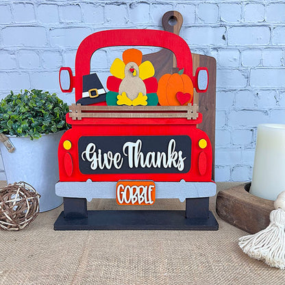 The "Give Thanks Interchangeable Insert - Painted" by Janet's Craft Corner features a decorative design of a red truck carrying a turkey, pumpkin, and pilgrim hat, adding charm to your home decor. The words "Give Thanks" appear on the truck, while "Gobble" is displayed on the license plate. Set against a brick wall with an adjacent plant and candle, it enhances any seasonal display.