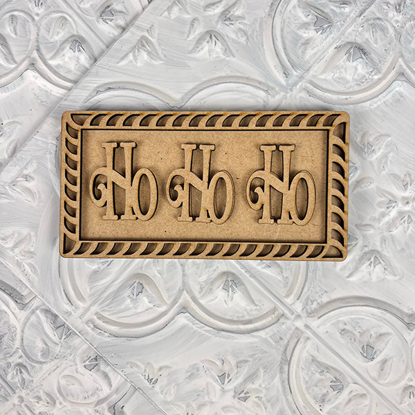 A rectangular wooden sign from Janet's Craft Corner, featuring the words "Ho Ho Ho" carved in bold letters, is centered on a textured white background. Ideal for your Vintage Santa Cookies Tiered Tray Set, this decorative piece boasts an ornate border that enhances the charm of your festive DIY Kit collection.