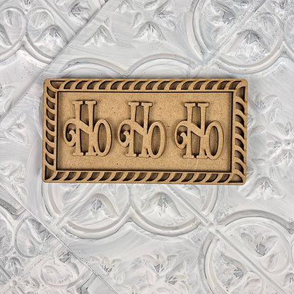 A rectangular wooden sign from Janet's Craft Corner, featuring the words "Ho Ho Ho" carved in bold letters, is centered on a textured white background. Ideal for your Vintage Santa Cookies Tiered Tray Set, this decorative piece boasts an ornate border that enhances the charm of your festive DIY Kit collection.