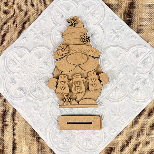 The Gingerbread Gnome Shelf Sitter by Janet's Craft Corner, featuring a festive hat and holding two gingerbread people, is adorned with snowflakes. Set against a white heart-patterned tile on burlap, this DIY seasonal decor craft kit adds charm to any festive setting.