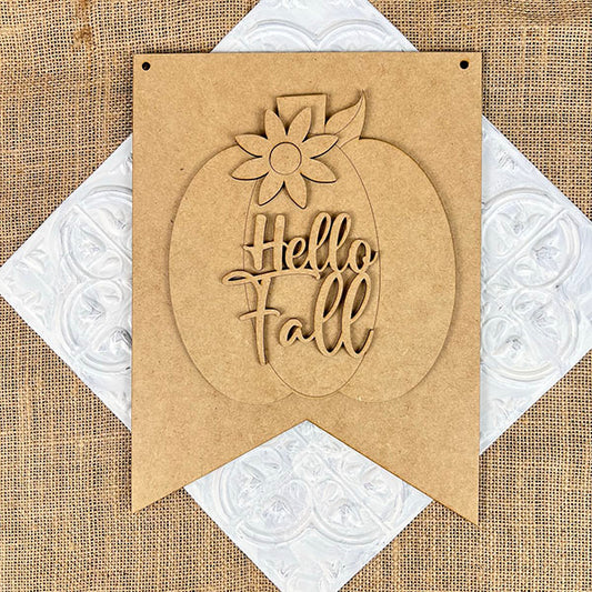 The "Hello Fall Wall Hanger" from Janet's Craft Corner features a carved pumpkin and flower atop "Hello Fall," ideal for seasonal décor on a textured white surface with a burlap background.