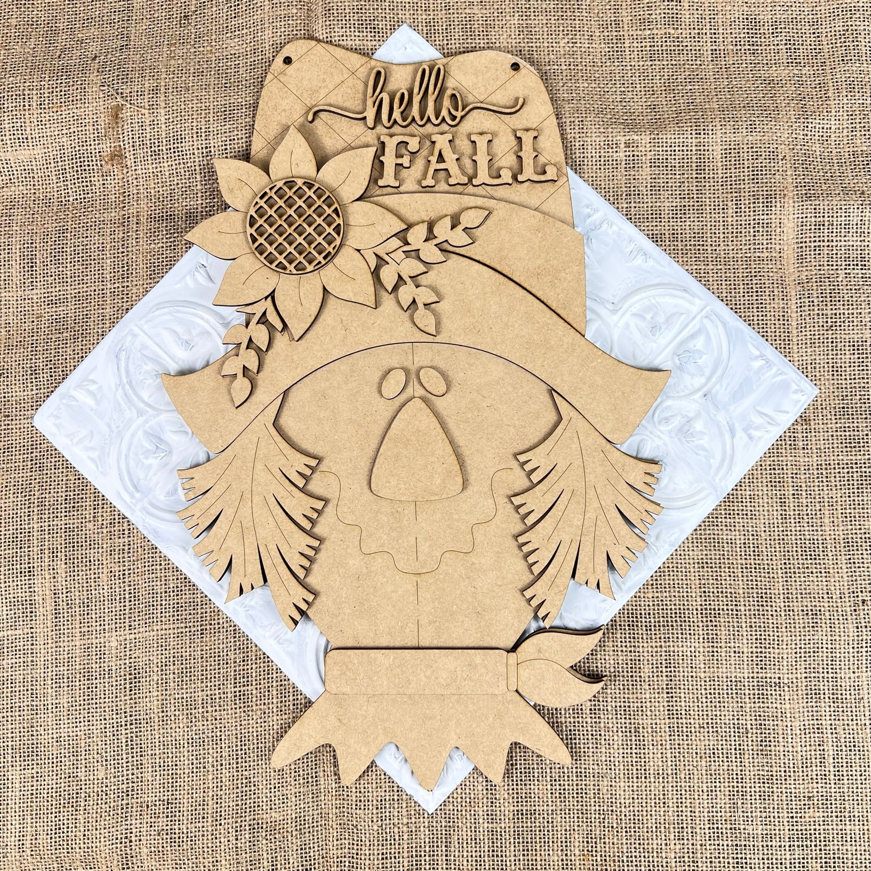 Janet's Craft Corner presents the Scarecrow Door Hanger, a wooden cutout shaped like a scarecrow face adorned with a sunflower on its hat and featuring the greeting "Hello Fall." Placed against a white textured diamond background on burlap, this charming piece adds an inviting touch to your home décor.