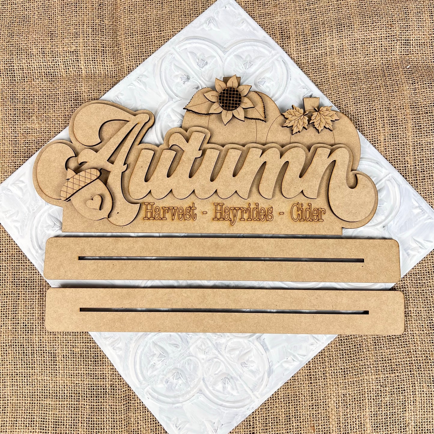 Introducing the Autumn Shelf Sitter from Janet's Craft Corner: This hand-painted wooden sign features ornate lettering with the word "Autumn" and is adorned with beautiful details such as pumpkins, a sunflower, acorns, and leaves. Additionally, it includes engraved words like "Harvest," "Hayrides," and "Cider." It serves as the perfect seasonal accent on a textured burlap background to complement your craft kits.