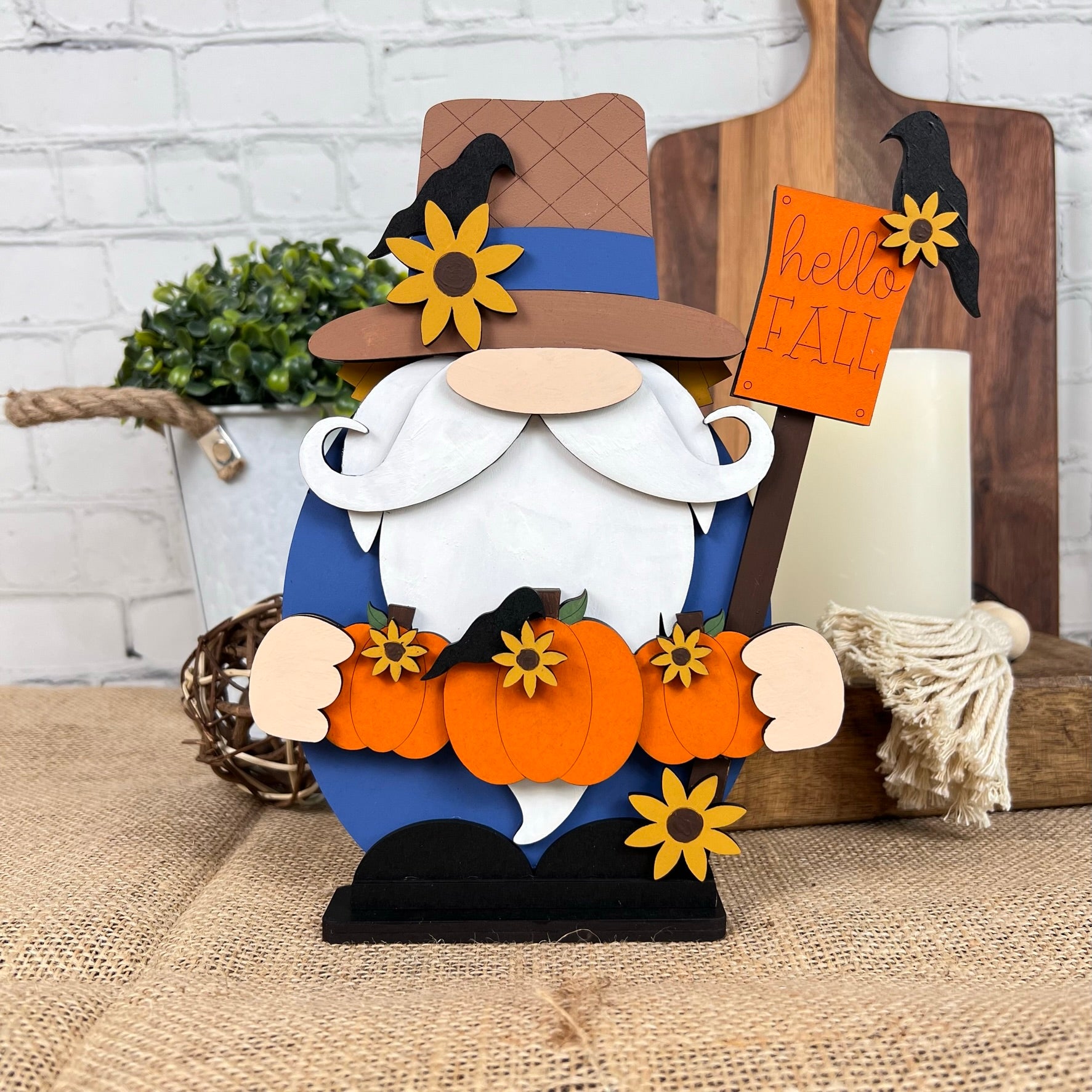 Introducing the Scarecrow Gnome from Janet's Craft Corner: a delightful wooden figurine featuring a whimsical design. This gnome boasts a long white beard and holds two vibrant orange pumpkins, while wearing a brown hat adorned with sunflowers. It carries a charming "hello FALL" sign, making it an ideal addition to your home décor. The rustic background enhances its seasonal charm, bringing cozy warmth to any space.