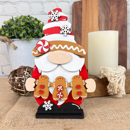 The Gingerbread Gnome DIY Kit by Janet's Craft Corner showcases a charming wooden Santa figure wearing a festive hat and accompanied by gingerbread men decorated with heart and candy motifs. The set includes additional decor elements such as a small plant, twine ball, candle, and wooden tray on a burlap surface against a brick wall backdrop, all of which create an instant holiday atmosphere.