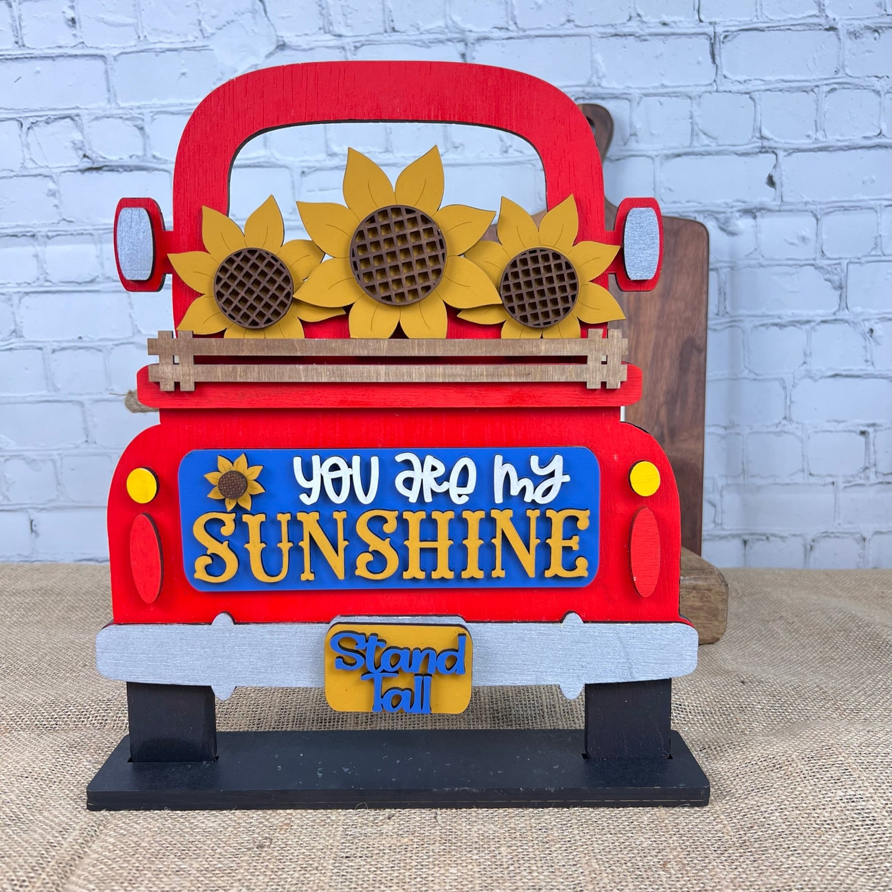 This delightful DIY home decor kit from Janet's Craft Corner features a red truck decorated with vibrant sunflowers. The message "You are my sunshine" stands out, complemented by a small placard that reads "Stand tall," all set against a rustic brick wall backdrop. Perfect for sunflower decor enthusiasts, this Sunflower Interchangeable Insert adds charm to any space.