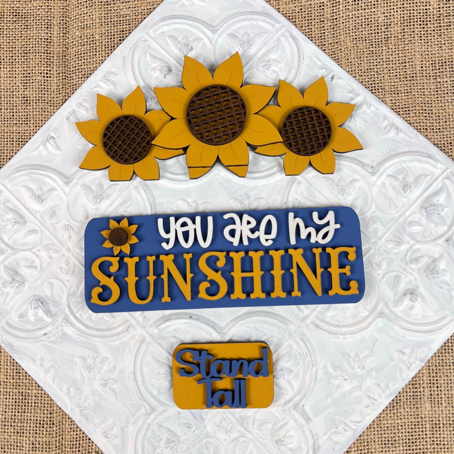 In the Sunflower Interchangeable Insert DIY home decor kit by Janet's Craft Corner, decorative cookies are beautifully set against a white embossed background, embodying the sunflower theme. Among these, three cookies are crafted into sunflower shapes; one is inscribed with "You are my sunshine" alongside a charming sunflower graphic, while another smaller one bears the message "Stand tall." The color palette features vibrant yellow, soothing blue, and earthy brown tones.