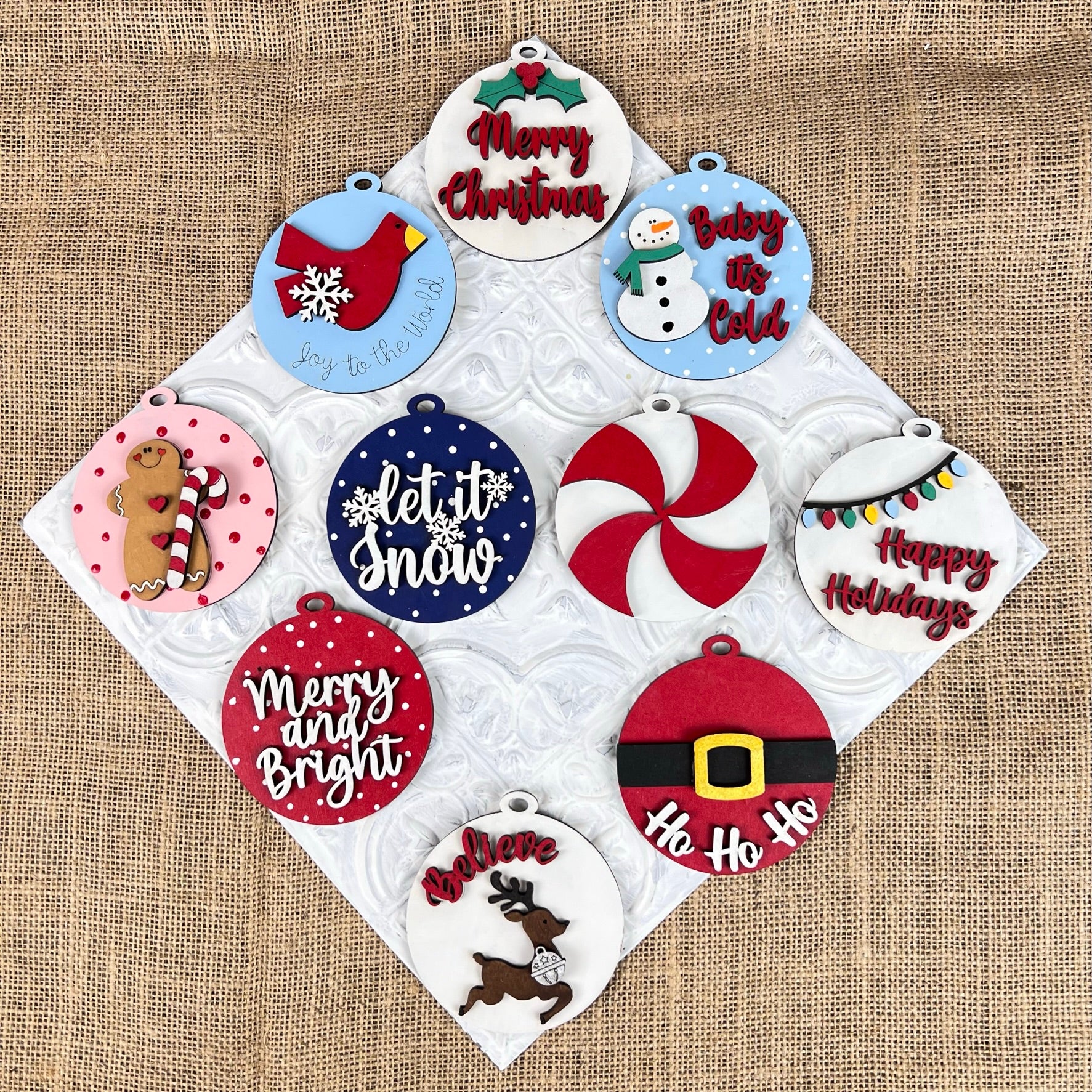 Janet's Craft Corner offers a DIY Decoration Kit featuring ten festive Christmas Cookies—round ornaments adorned with red and white patterns, snowmen, reindeer, Christmas trees, candy cane swirls, and holiday messages like "Merry Christmas" and "Happy Holidays." These beautifully designed cookies can be artistically arranged among your holiday decorations on a burlap background.