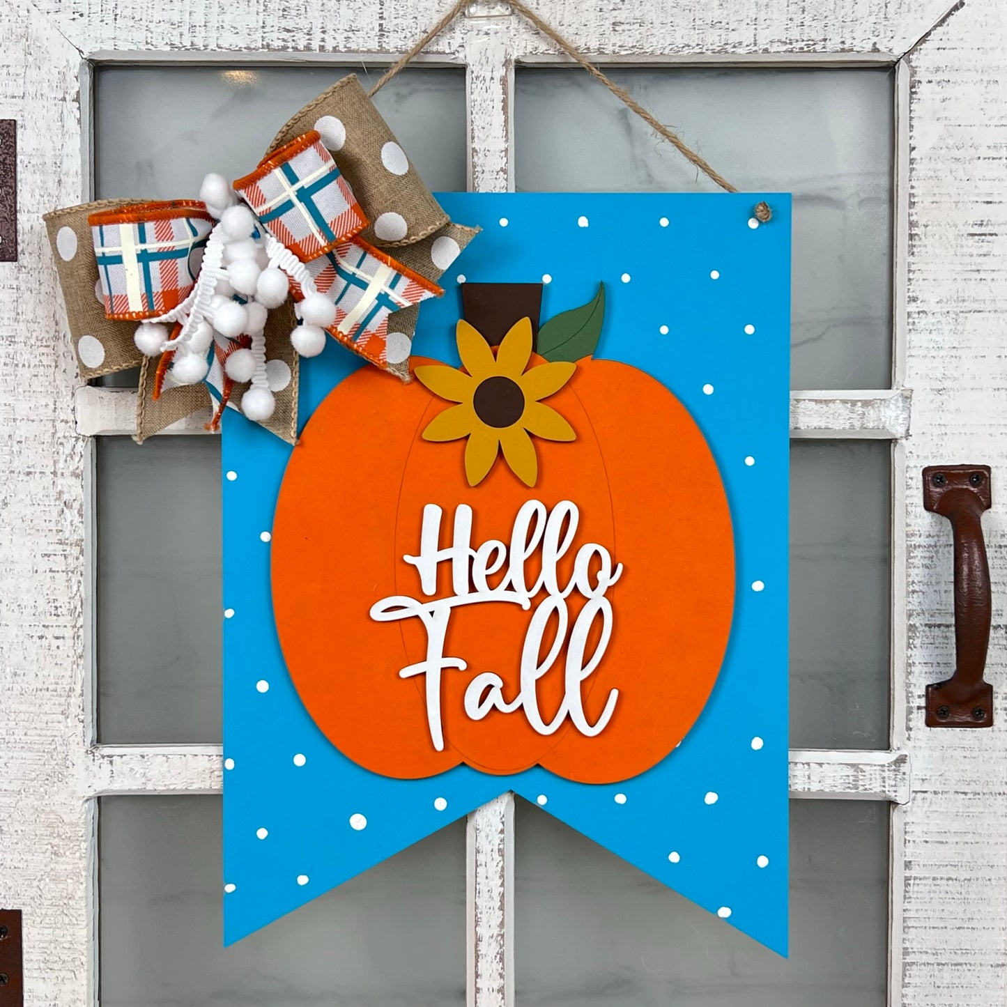 The "Hello Fall Wall Hanger" from Janet's Craft Corner is a handcrafted decoration featuring an orange pumpkin and a sunflower set against a blue background with white dots on a white door. With the text "Hello Fall," it's adorned with a large plaid and pom-pom bow, adding charm to any entrance this autumn.