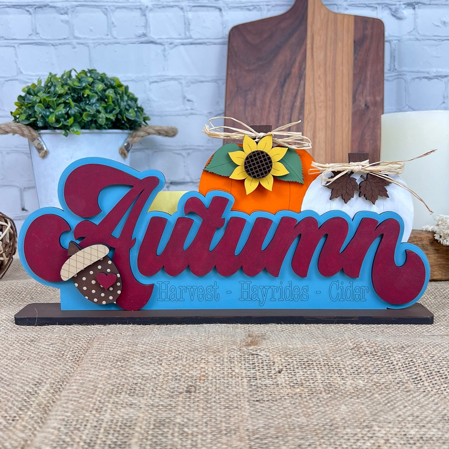 Janet's Craft Corner presents the Autumn Shelf Sitter, a hand-painted wooden sign that spells "Autumn" in red. The design is completed with autumn-themed decorations such as a sunflower, acorns, leaves, and a pumpkin. The words "Harvest," "Hayrides," and "Cider" are beautifully etched below. This charming piece is perfect as a shelf sitter and is displayed against chopping boards with a small potted plant adding to the seasonal ambiance.