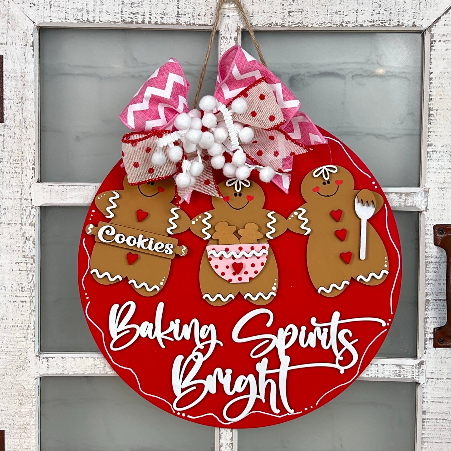 The "Baking Spirits Bright Door Hanger" from Janet's Craft Corner showcases a lively red backdrop adorned with three gingerbread figures dressed in holiday outfits. The phrase "Baking Spirits Bright" is elegantly scripted in white, accompanied by delightful accents of red, pink, and white ribbons and cheerful pom-poms, embodying the spirit of joyful home décor.