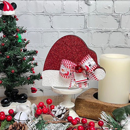 A festive scene features a small Christmas tree and Janet's Craft Corner's Chunky Santa Hat decoration, adorned with a striped bow. Accompanied by a lit candle and red berries on the table, this holiday decor set against a white brick wall brings cozy charm to any space.