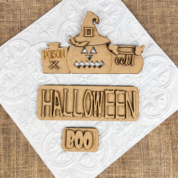 Janet's Craft Corner presents the Halloween Pumpkin Interchangeable Insert—an ideal DIY home decor kit. It features wooden Halloween decorations on a textured white background: a pumpkin adorned with a witch hat labeled "poison" and "eek!", alongside a sign that reads "HALLOWEEN," and a small plaque with the word "BOO!" All elements rest atop burlap fabric, offering perfect pieces for interchangeable craft inserts.