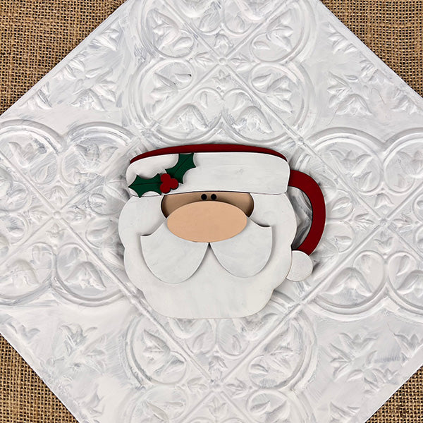 A piece from the Vintage Santa Cookies Tiered Tray Set by Janet's Craft Corner features a wooden Santa Claus face decoration resting on a white embossed background. The Santa is adorned with a red hat, green holly leaf accents, and a red nose, reminiscent of classic vintage Santa cookies.