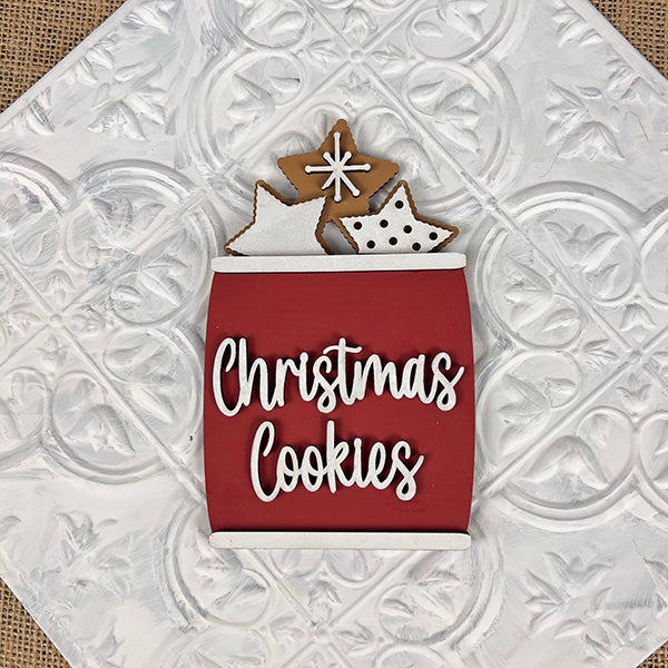 A red container labeled "Vintage Santa Cookies Tiered Tray Set" by Janet's Craft Corner is filled with gingerbread stars, some adorned with white icing and dots, capturing the nostalgic charm of Christmas cookies. The setting includes an embossed white tile pattern in the background, making it perfect for a festive display.