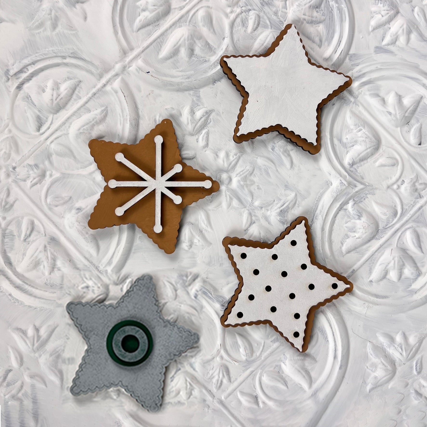 Four cookies from Janet's Craft Corner's Vintage Santa Cookies Tiered Tray Set sit on a white textured background. Shaped like stars and adorned with icing designs, one cookie is decorated with a snowflake, another remains plain white, the third features black dots, and the fourth is gray with a circular center.