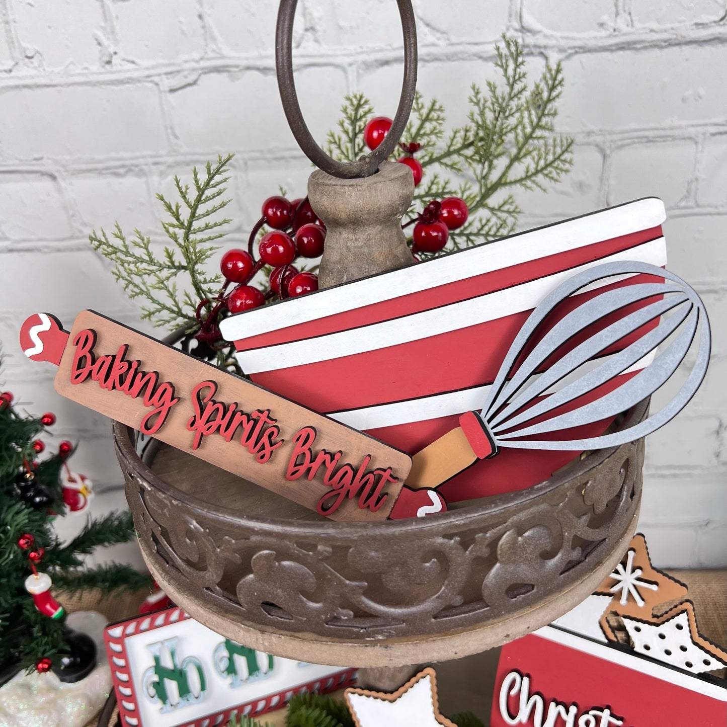 Introducing the Vintage Santa Cookies Tiered Tray Set from Janet's Craft Corner, featuring a charming metal stand decorated with holiday-themed decor elements. This festive display includes a whisk and a sign with the cheerful message "Baking Spirits Bright," all beautifully accented by red and white stripes, faux greenery embellished with red berries, and whimsical touches of vintage Santa cookies.