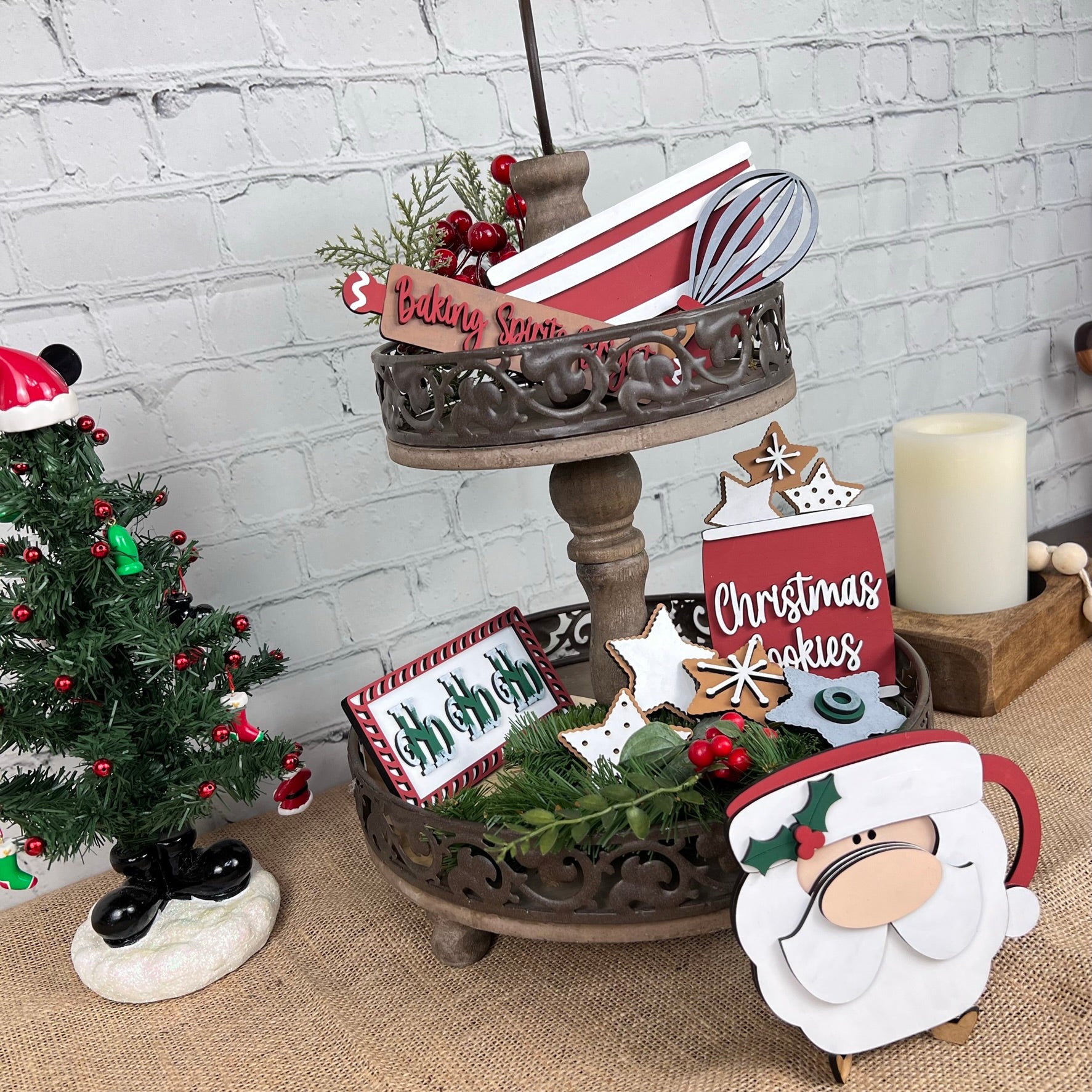 The Vintage Santa Cookies Tiered Tray Set by Janet's Craft Corner highlights holiday decorations with charming small signs such as "Baking Spirits Bright" and "Christmas Cookies," all accented by greenery and berries. It lends a whimsical feel with nearby Vintage Santa Cookies, complemented by a small decorated Christmas tree on a burlap surface.