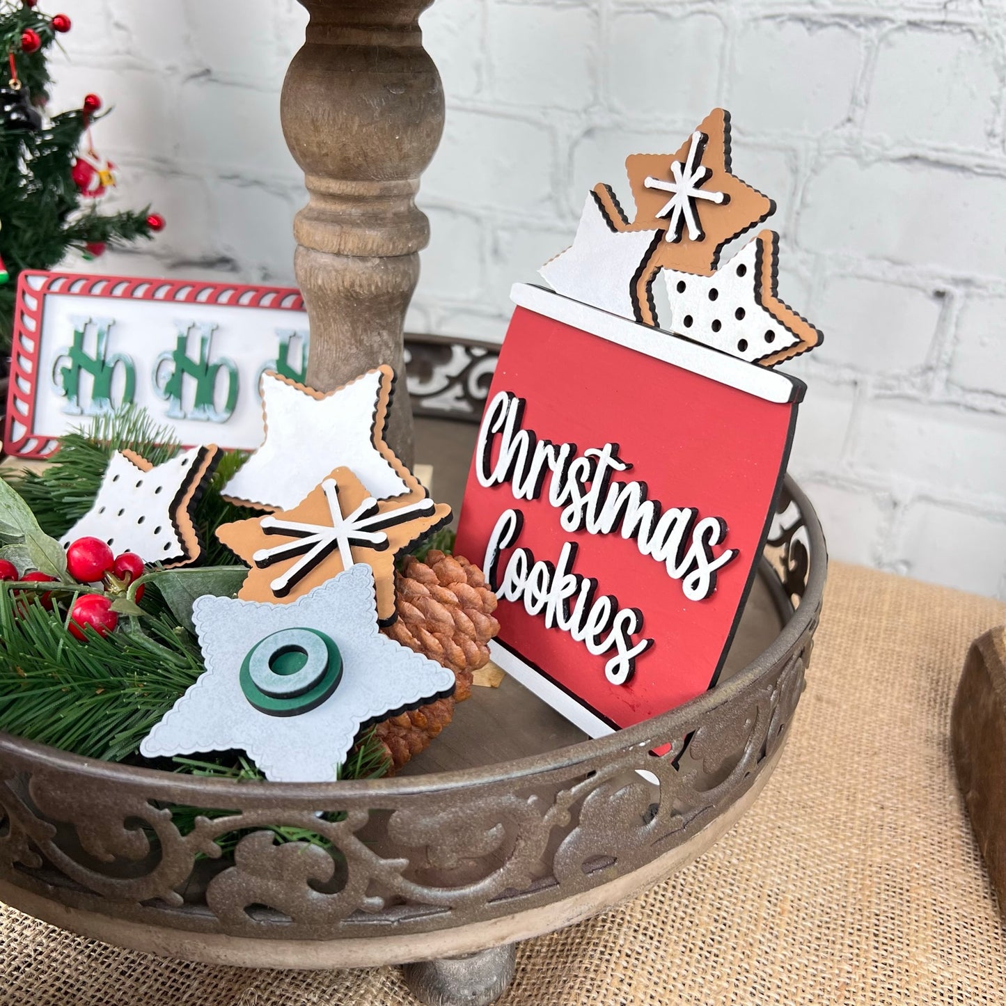 A decorative display showcases the "Christmas Cookies" red sign from Janet's Craft Corner's Vintage Santa Cookies Tiered Tray Set. The scene is adorned with star-shaped cookies, some topped with icing and sprinkles, all nestled on a tiered tray set. Festive greenery and a small Christmas tree complete the background ambiance.