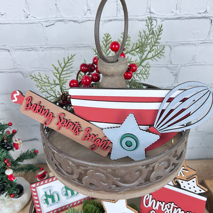 A decorative display showcases the Vintage Santa Cookies Tiered Tray Set from Janet's Craft Corner, featuring a red and white striped bowl, a wooden whisk, and a sign that reads "Baking Spirits Bright." The arrangement is accentuated with greenery, red berries, Vintage Santa cookies, and Christmas-themed ornaments on a rustic background.