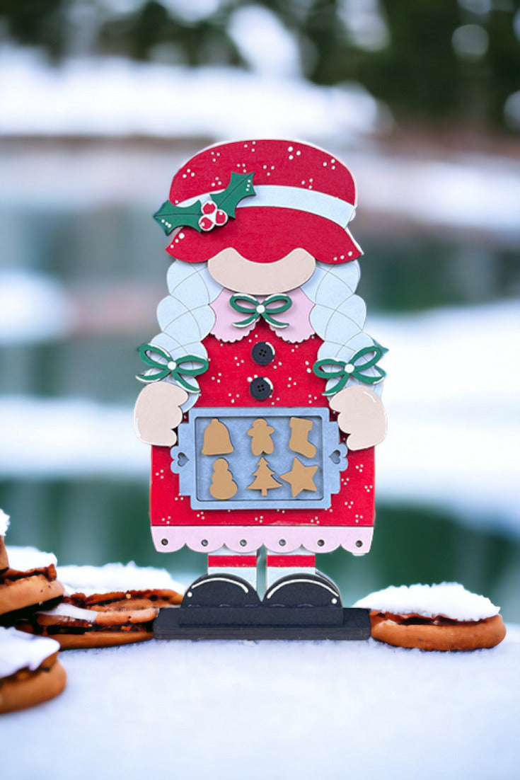 Decorate your home with Janet's Craft Corner's Mrs Claus Baking Gnome Shelf Sitter. This DIY seasonal décor kit includes a Mrs Claus in a red dress holding holiday cookies, set against a snowy background with blurred greenery and cookies stacked in the foreground.