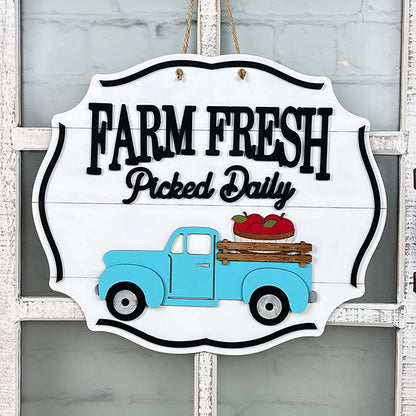 A rustic Farm Fresh Apples Sign by Janet's Craft Corner, featuring the bold black phrase "Farm Fresh Picked Daily" alongside a charming illustration of a blue vintage truck carrying red apples, is beautifully set against a white background.