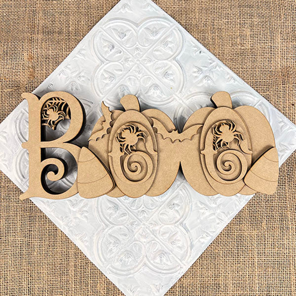 The Chunky Boo Shelf Sitter by Janet's Craft Corner spells "BOO" with scrollwork, featuring two pumpkins and leaves. This DIY seasonal home decor piece rests angled on a white textured square over burlap, perfect for Halloween.