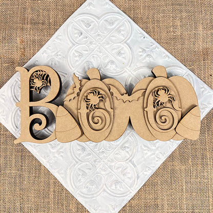 The Chunky Boo Shelf Sitter by Janet's Craft Corner spells "BOO" with scrollwork, featuring two pumpkins and leaves. This DIY seasonal home decor piece rests angled on a white textured square over burlap, perfect for Halloween.