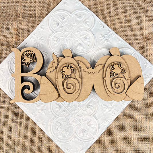 The Chunky Boo Shelf Sitter by Janet's Craft Corner spells "BOO" with scrollwork, featuring two pumpkins and leaves. This DIY seasonal home decor piece rests angled on a white textured square over burlap, perfect for Halloween.