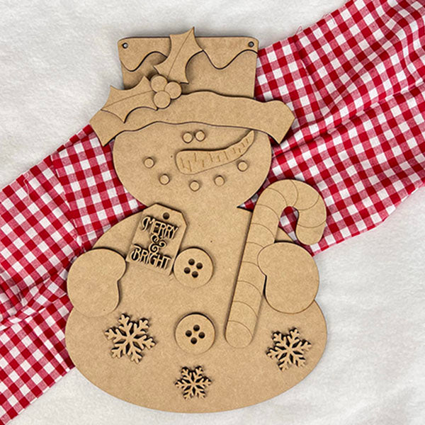 The Candy Cane Snowman Door Hanger from Janet's Craft Corner is a delightful wooden decoration on a red and white checkered cloth, featuring button and snowflake cutouts. Holding a candy cane, it wears a holly-adorned top hat and displays a "Merry & Bright" tag, perfect for holiday decor lovers.