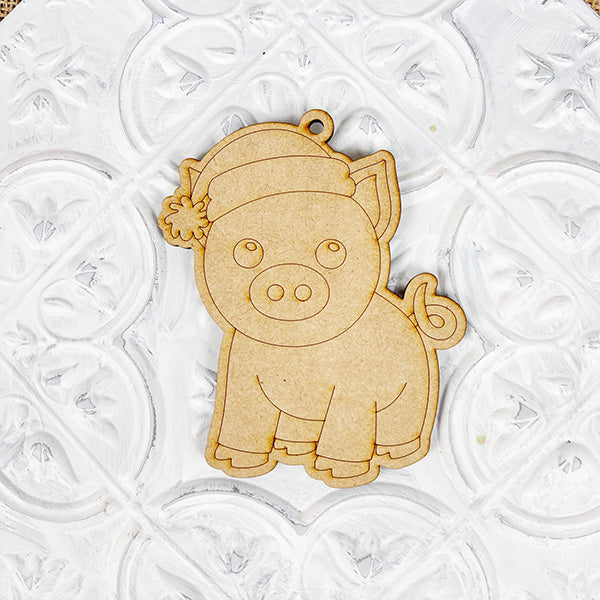 The Color Your Own Ornament kit from Janet's Craft Corner features a charming wooden pig in a Santa hat against a patterned white backdrop, ideal for craft lovers interested in home décor. With detailed engravings and a hanging loop, this DIY Decoration Kit invites you to personalize it with your creative flair.