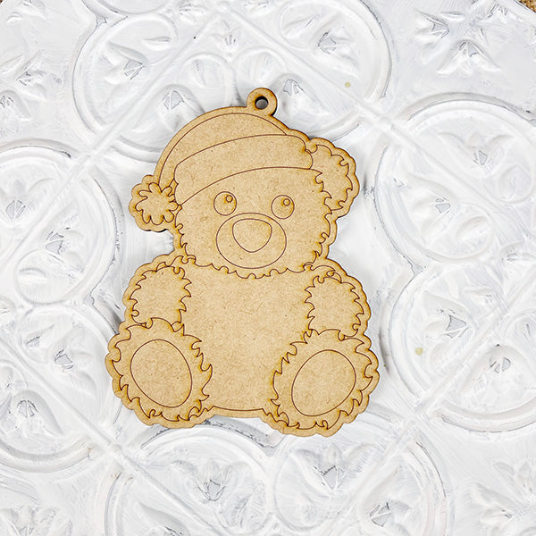 A wooden cutout of a teddy bear wearing a Santa hat rests on a white textured surface, making it an ideal choice for fans of Janet's Craft Corner's Color Your Own Ornament | DIY Decoration Kits. The charming and straightforward design includes etched details that outline its features, encouraging you to use your creativity to color the ornament and personalize your home décor.